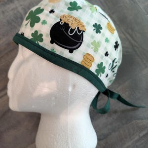 Op de afbeelding: Een wit en groen patroon scrub cap met een zwarte pot met goud, klavers en gouden munten. De cap heeft een groene bies en strikbanden. De stof heeft een ruitpatroon.