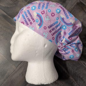 May include: A light purple scrub cap with a repeating pattern of rainbows, smiley faces, and flowers. The text "Sarcasm is my first language" is printed on the cap in a playful font. The cap is designed to be worn in a medical or professional setting.