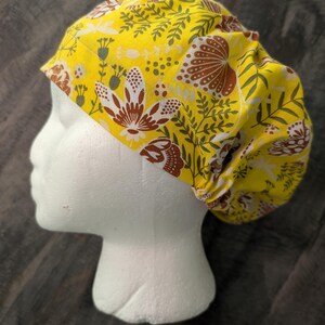 Puede incluir: Un gorro quirúrgico amarillo con un estampado floral. El gorro presenta un diseño repetido de flores marrones y blancas y hojas verdes sobre un fondo amarillo. El gorro está diseñado para cubrir la cabeza y el cabello.