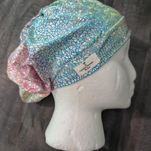 May include: A scrub cap featuring a holographic, iridescent, cracked-glass pattern in shades of blue, green, and pink. The cap has a gathered back and a white fabric label on the side that reads Carolina Dreams By Jen with a palm tree icon.