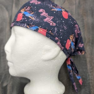 Pode incluir: Um gorro cirúrgico azul escuro com uma estampa de fada rosa e azul. O gorro tem um laço na parte de trás e o texto "The Tonsil Fairy" está impresso na parte da frente.