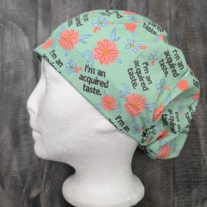 Pode incluir: Uma touca cirúrgica verde clara com um padrão floral de flores rosa e azuis. A touca tem o texto "I'm an acquired taste" impresso. A touca está em uma cabeça de manequim branca.
