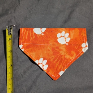 May include: Orange and white dog bandana with paw print pattern. The bandana is approximately 15 cm long. The fabric has a tie-dye effect. The bandana is made of fabric and has a white stitched edge.