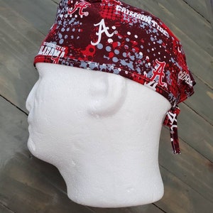 May include: A burgundy and red scrub cap featuring the University of Alabama's logo and the words "Crimson Tide." The cap has a tie in the back and is displayed on a white mannequin head.