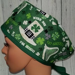 Könnte beinhalten: Grün-weiß gemusterte OP-Haube mit St. Patrick's Day-Motiv. Die Haube zeigt Kleeblätter, einen Koboldhut und die Aufschriften "Kiss Me", "I'm Irish!" und "Go Green". Die Haube hat einen grünen Rand und Bindebänder.