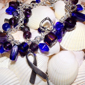 K&ouml;nnte beinhalten: Ein silbernes Armband mit lila und blauen Glasperlen, einem silbernen Herz-Anh&auml;nger und einem silbernen Band-Anh&auml;nger. Das Armband liegt auf einem Bett aus wei&szlig;en Muscheln.