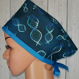 Op de afbeelding: Een blauwe chirurgische scrub-muts met een wit DNA-patroon en een turquoise blauwe rand. De muts heeft banden aan de achterkant.