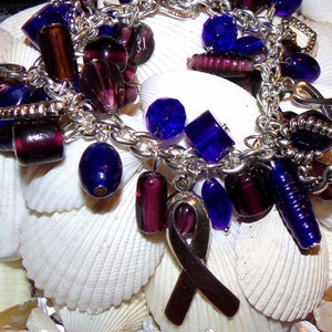 K&ouml;nnte beinhalten: Ein silbernes Armband mit lila und blauen Glasperlen, einem silbernen Band-Anh&auml;nger und anderen silbernen Anh&auml;ngern. Das Armband wird auf einem Bett aus wei&szlig;en Muscheln pr&auml;sentiert.