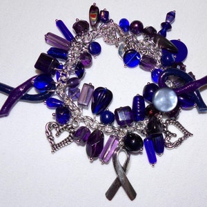 K&ouml;nnte beinhalten: Ein silbernes Armband mit violetten und blauen Glasperlen, Charms und einem violetten Band. Das Armband besteht aus einer silbernen Kette und hat eine Vielzahl von Charms, darunter Herzen, ein Band und eine kleine violette Blume.