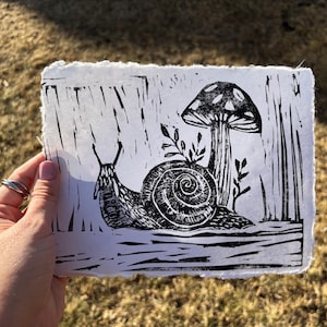 May include: Black and white linocut print featuring a snail, mushroom, and foliage. The artwork has a rustic, handmade aesthetic with visible texture and a deckled edge. The print is held against a blurred outdoor background.