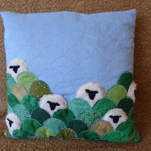 May include: A blue pillow with a quilted design of overlapping semicircles. The pillow is decorated with felt sheep appliques in a row along the bottom edge. The sheep are white with black faces and are surrounded by green felt patches that resemble hills.