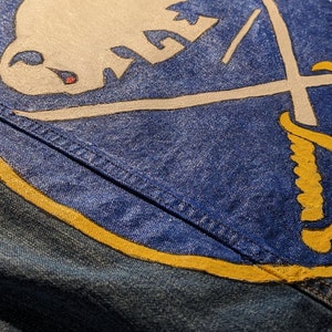 May include: Close-up of a denim jacket featuring a large, hand-painted Buffalo Sabres logo in white, blue, and yellow. The logo includes a charging buffalo and crossed swords. The word "BUFFALO" is painted in yellow.