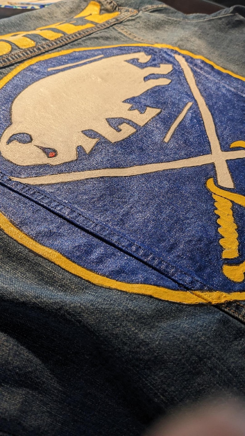 May include: Close-up of a denim jacket featuring a large, hand-painted Buffalo Sabres logo in white, blue, and yellow. The logo includes a charging buffalo and crossed swords. The word "BUFFALO" is painted in yellow.