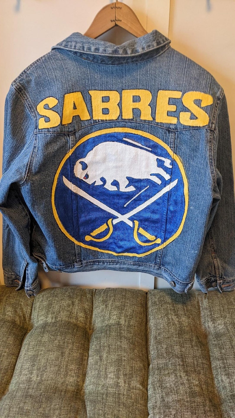 May include: A denim jacket with the word "SABRES" in yellow block letters across the back. Below the text is a circular emblem featuring a white buffalo, crossed swords, and a yellow border, all on a blue background.