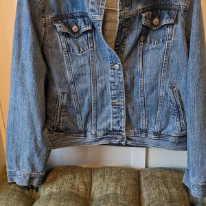 May include: A medium-wash denim jacket with a classic collar, button-down front, and two chest pockets. The jacket is hanging on a wooden hanger. The jacket is displayed on a green tufted cushion.