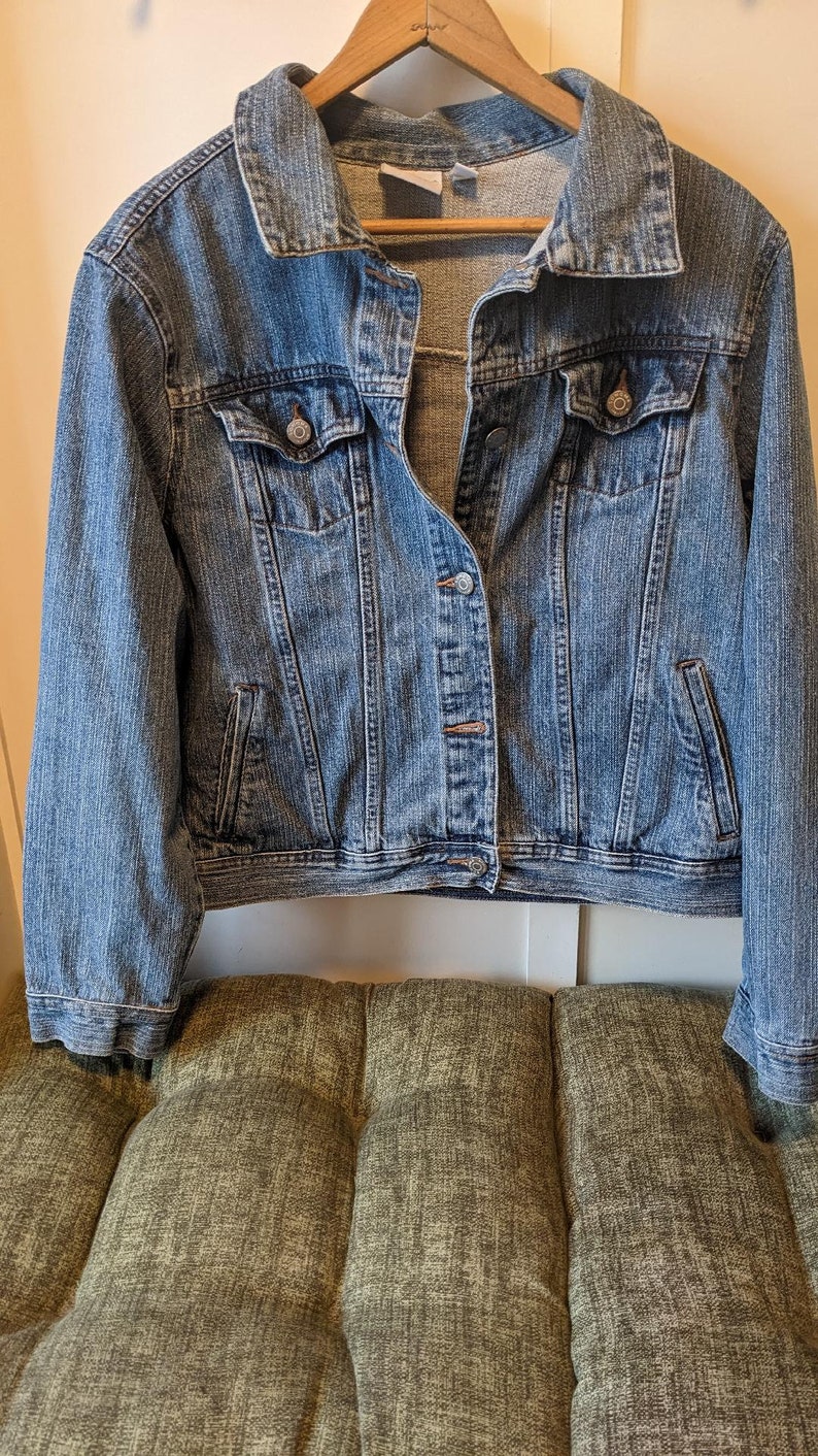 May include: A medium-wash denim jacket with a classic collar, button-down front, and two chest pockets. The jacket is hanging on a wooden hanger. The jacket is displayed on a green tufted cushion.