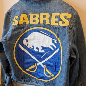 May include: A denim jacket with the word "SABRES" in yellow block letters across the shoulders. The back of the jacket features a blue and yellow logo with a white bison and crossed swords, representing the Buffalo Sabres.