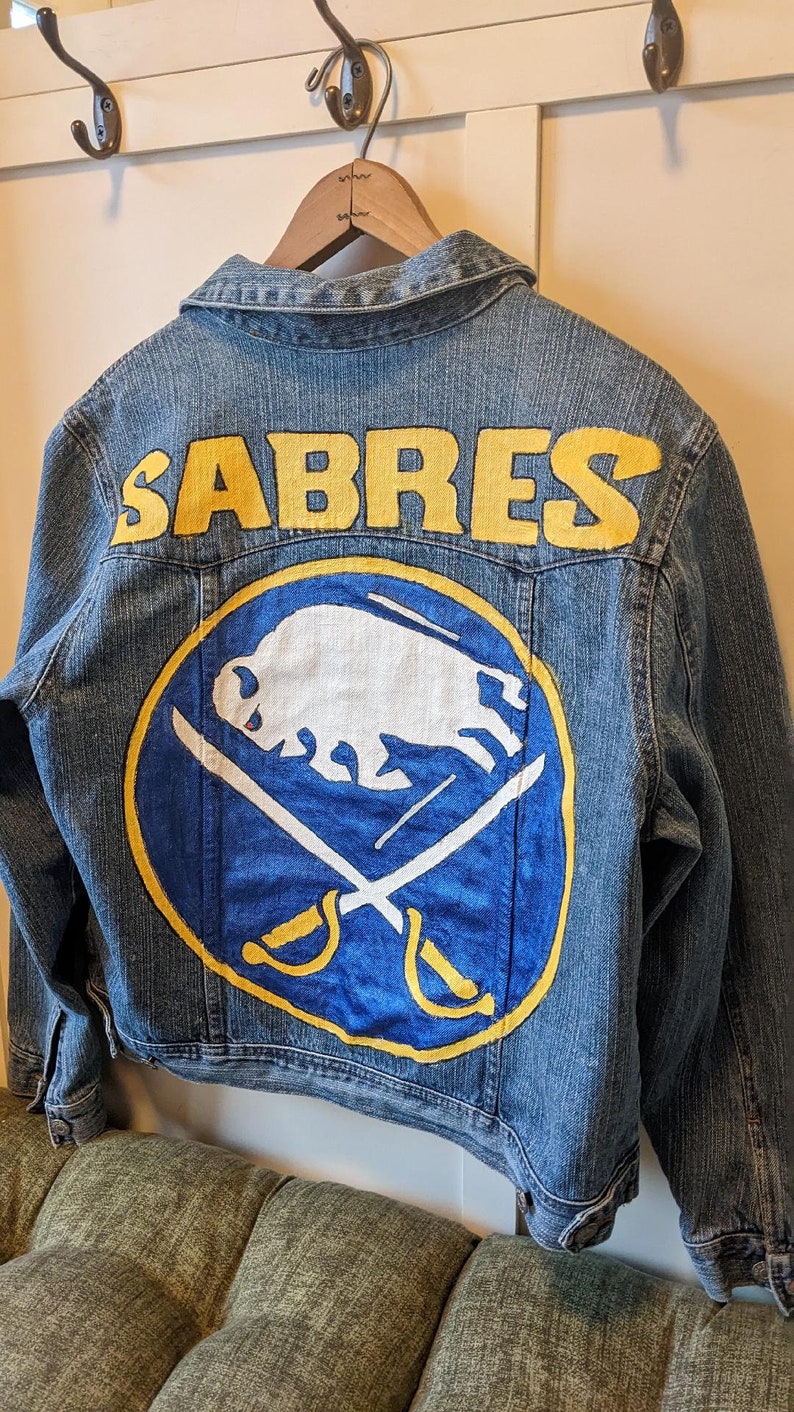 May include: A denim jacket with the word "SABRES" in yellow block letters across the shoulders. The back of the jacket features a blue and yellow logo with a white bison and crossed swords, representing the Buffalo Sabres.
