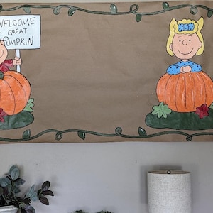 Może przedstawiać: Brązowy papierowy baner z postaciami z komiksu Peanuts. Charlie Brown trzyma znak z napisem "Welcome Great Pumpkin". Sally Brown jest r&oacute;wnież obecna. Baner jest ozdobiony zieloną obw&oacute;dką z winorośli i pomarańczowymi dyniami.