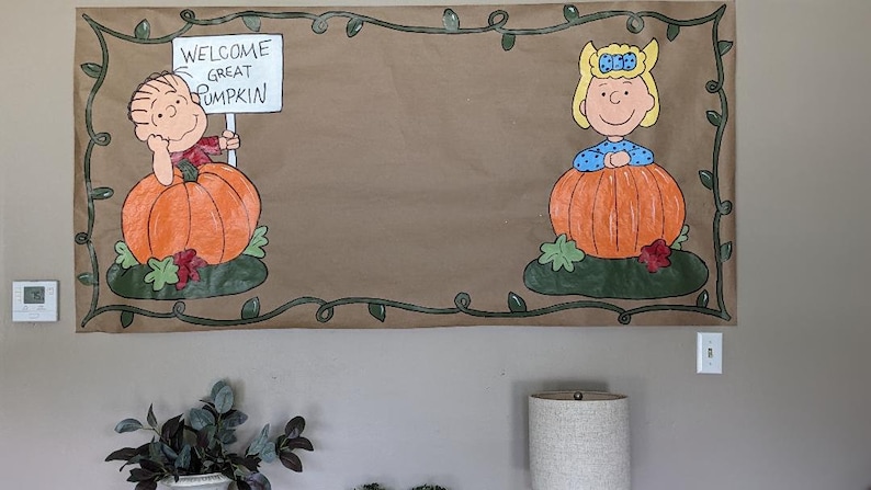 Może przedstawiać: Brązowy papierowy baner z postaciami z komiksu Peanuts. Charlie Brown trzyma znak z napisem "Welcome Great Pumpkin". Sally Brown jest r&oacute;wnież obecna. Baner jest ozdobiony zieloną obw&oacute;dką z winorośli i pomarańczowymi dyniami.