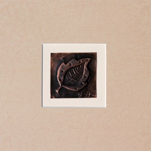 May include: A framed copper leaf sculpture with a textured surface and a raised design. The leaf is brown and has a small inscription that reads "kade".
