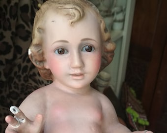 May include: A vintage porcelain baby doll with painted features, including rosy cheeks and brown eyes. The doll has light brown curly hair and is wearing a pair of off-white shorts. The doll is being held in a person's hand.