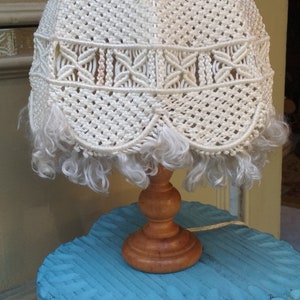 Op de afbeelding: Een witte macram&eacute; lampenkap met een franje rand op een blauwe houten basis. De lampenkap heeft een uniek ontwerp met ingewikkelde knopen en een gegolfde rand.