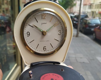 Könnte beinhalten: Ein weißer Vintage-Wecker mit einem goldenen Zifferblatt, auf dem "Goldblatt Musical Alarm" steht. Der Wecker hat einen kleinen Plattenspieler an der Unterseite, mit einer schwarzen Schallplatte, auf der "Alte Skaters - Waltz" steht.