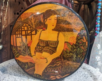 Può includere: Contenitore decorativo rotondo e vintage con un'immagine dipinta di una donna in un paesaggio. L'opera d'arte presenta toni caldi di arancione, marrone e giallo. La donna indossa un abito fantasia. Il contenitore ha un bordo nero.