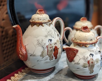 May include: A set of three white porcelain teapots with red and gold accents. The teapots are decorated with a Japanese-inspired floral design and feature a scene of women in traditional clothing.