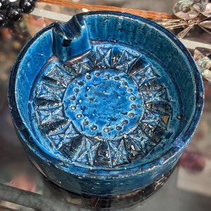Puede incluir: Un cenicero de cerámica azul con un diseño geométrico. El cenicero tiene un borde elevado y una pequeña hendidura para cigarrillos.