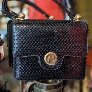 Può includere: Una borsa a mano in pelle di serpente nera con chiusura dorata e manico superiore. La borsa ha una superficie testurizzata e squamosa.