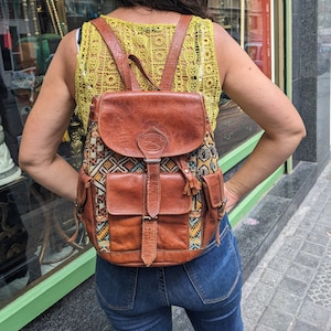 Puede incluir: Una mochila de cuero marrón con una solapa estampada de colores y un bolsillo frontal grande. La mochila tiene un cierre de hebilla y un borla.