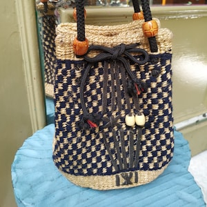 Puede incluir: Un pequeño bolso tejido con un patrón de cuadros blanco y negro. El bolso tiene un cierre de cordón y cuentas de madera en las asas. El bolso está hecho de fibras naturales y tiene un aspecto rústico.