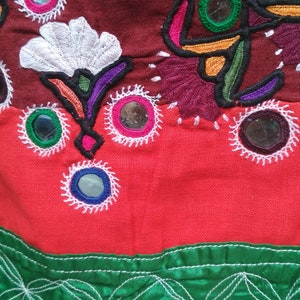 May include: A close-up of a red, green, and yellow fabric with intricate embroidery and small round mirrors. The fabric has a white floral design and a border of green and yellow.