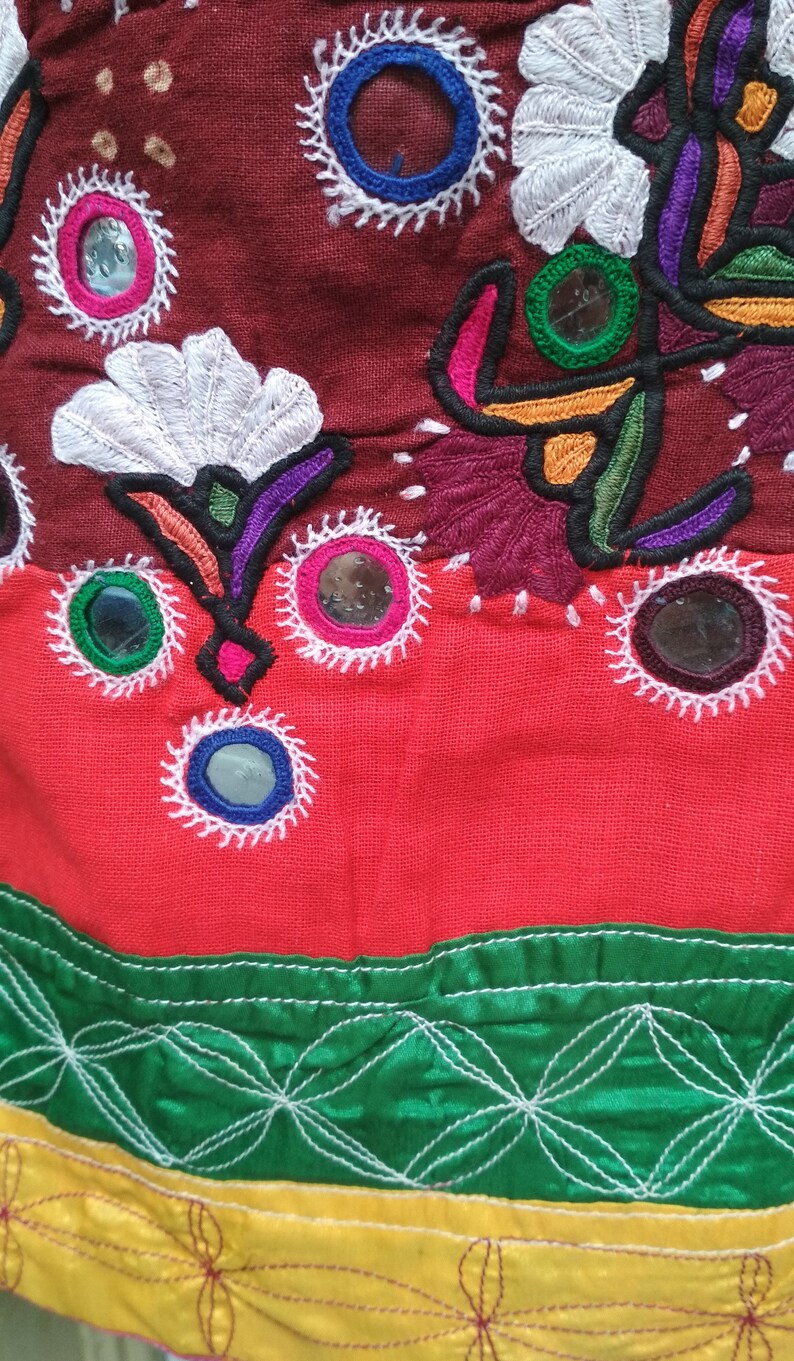 May include: A close-up of a red, green, and yellow fabric with intricate embroidery and small round mirrors. The fabric has a white floral design and a border of green and yellow.