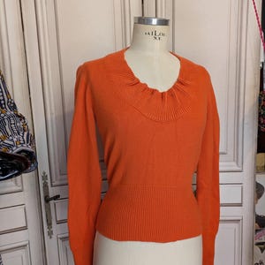 May include: An orange long-sleeved sweater on a mannequin. The sweater features a round neckline with a ruffled detail and a ribbed waist. The mannequin is labelled "TAILOR N.Y."