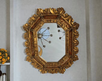 Könnte beinhalten: Achteckiger Spiegel mit einem goldenen, verzierten Rahmen. Der Rahmen hat ein detailliertes, gerolltes Design mit rotem Akzent. Der Spiegel reflektiert einen Kronleuchter und andere Objekte. Er ist an einer hellgrauen Wand befestigt.