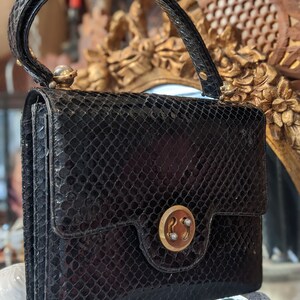 Puede incluir: Un bolso de mano de piel de serpiente negra con cierre dorado y una sola perla en el asa.