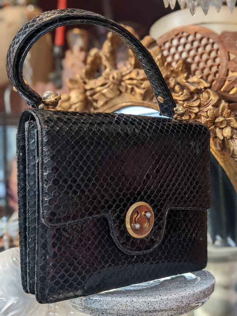 Puede incluir: Un bolso de mano de piel de serpiente negra con cierre dorado y una sola perla en el asa.