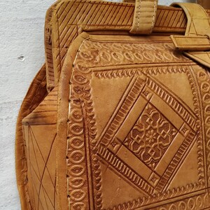 Puede incluir: Un bolso de mano de cuero marr&oacute;n con intrincados patrones en relieve y costuras. El bolso tiene un asa superior y un cierre de solapa.