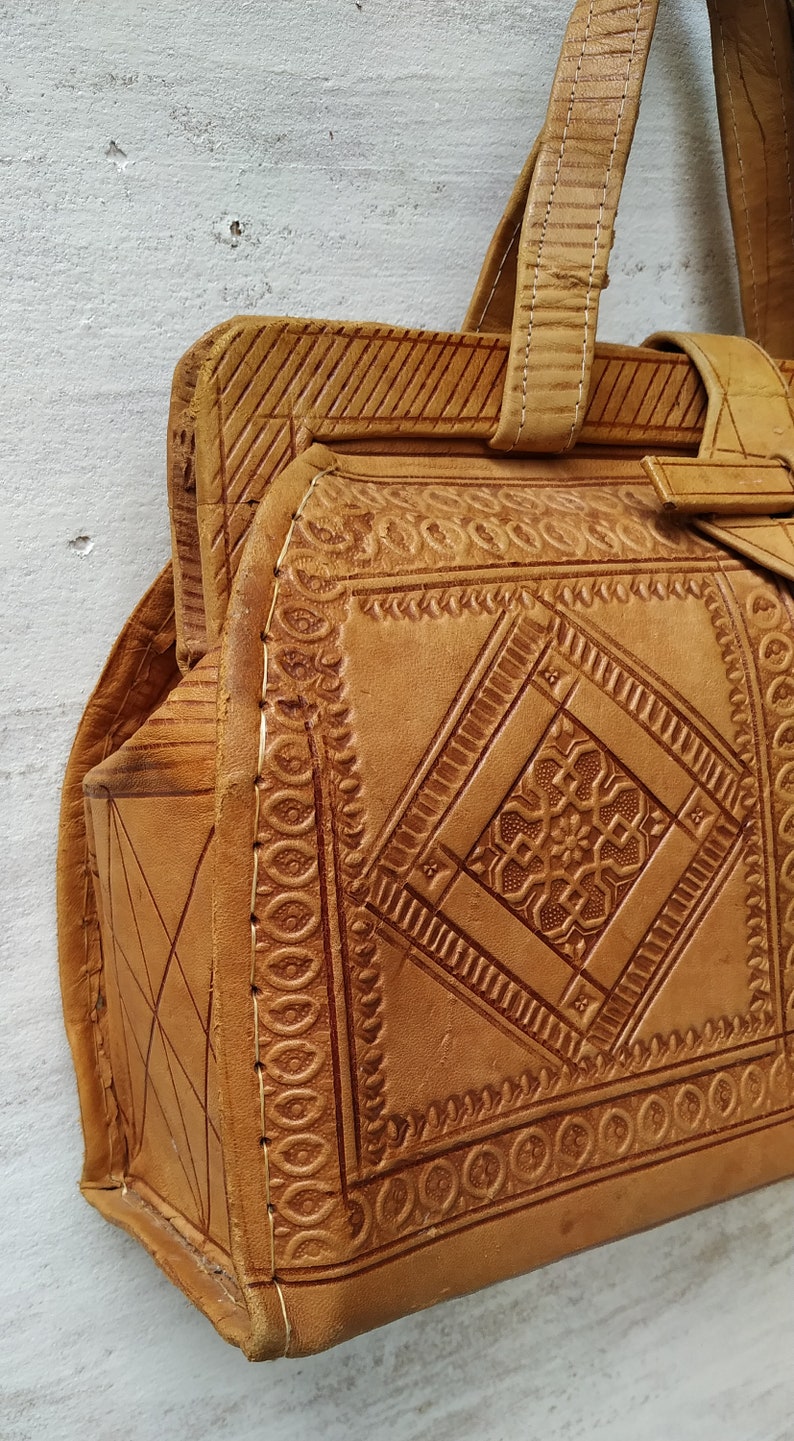 Puede incluir: Un bolso de mano de cuero marr&oacute;n con intrincados patrones en relieve y costuras. El bolso tiene un asa superior y un cierre de solapa.