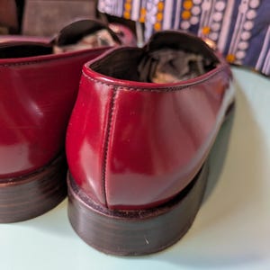 May include: Close-up of two burgundy leather loafers. The shoes have a glossy finish and dark brown soles. The stitching is visible along the seams. The loafers are a classic style.
