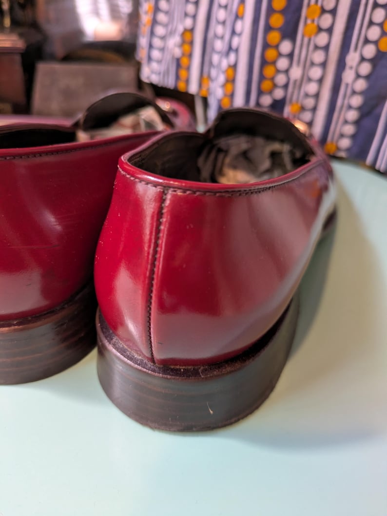 May include: Close-up of two burgundy leather loafers. The shoes have a glossy finish and dark brown soles. The stitching is visible along the seams. The loafers are a classic style.
