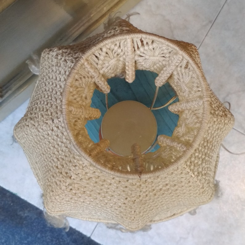 Op de afbeelding: Een beige gehaakte lampenkap met een ronde, open bovenkant. De lampenkap is gemaakt van een dik, getextureerd garen en heeft een gefranst randje.