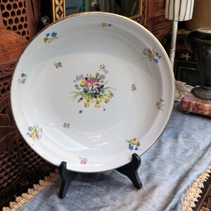 May include: A white ceramic bowl with a gold rim and a floral design. The bowl is decorated with small, colorful flowers and a larger bouquet of flowers in the center. The bowl is sitting on a black stand.
