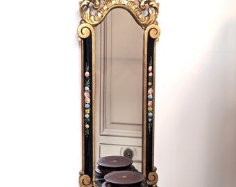 May include: A gold-framed wall mirror with a black background and floral accents. The mirror has a shelf with two round wooden platforms.