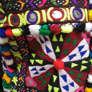 May include: A colorful textile with intricate embroidery, featuring geometric patterns, mirrors, and pom-poms. The fabric is a mix of red, green, blue, yellow, and white.