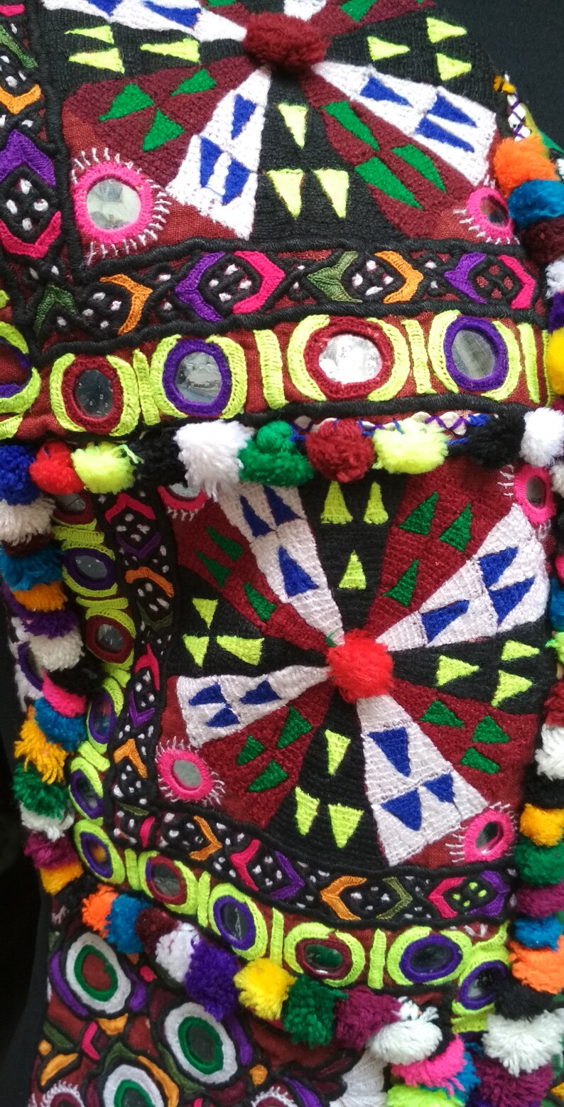 May include: A colorful textile with intricate embroidery, featuring geometric patterns, mirrors, and pom-poms. The fabric is a mix of red, green, blue, yellow, and white.