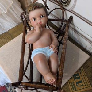 May include: A vintage doll with a painted face and light blue diaper sits in a rustic wooden cradle. The doll has brown hair and is posed with a finger in its mouth. The cradle is made of thin, intertwined branches.
